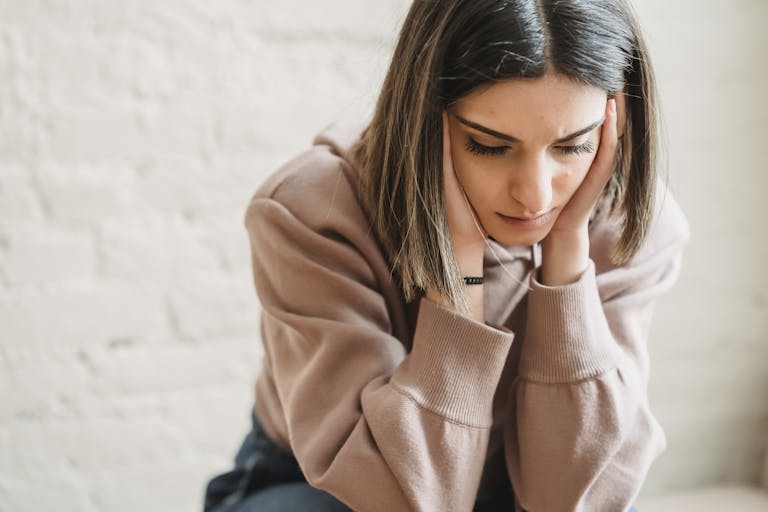 Crop unhappy female in casual wear touching cheeks and looking down while sitting near white wall in light room at home waiting for her chronic illness diagnosis